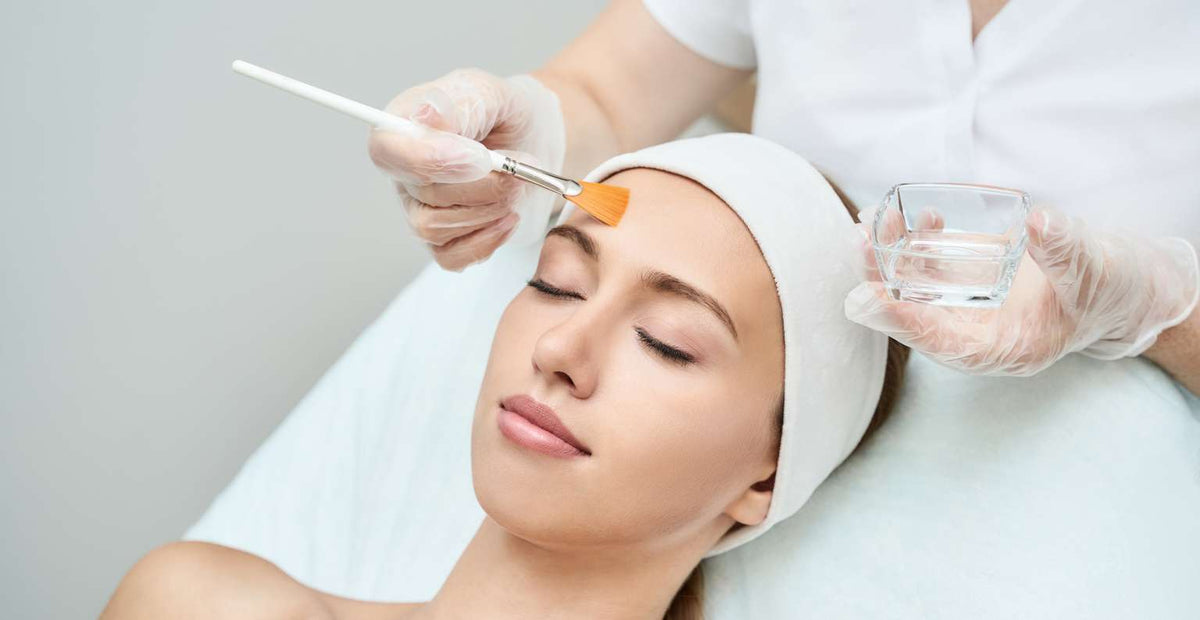 A woman with a white head wrap receives a facial treatment, with a gloved hand applying orange-colored cream to her forehead using a brush while another hand holds a small glass bowl.