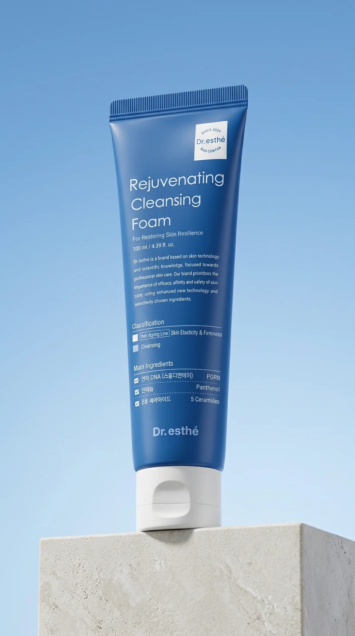 A blue squeeze tube of Dr. esthé Rejuvenating Cleansing Foam stands upright on a light stone pedestal against a soft blue gradient background.